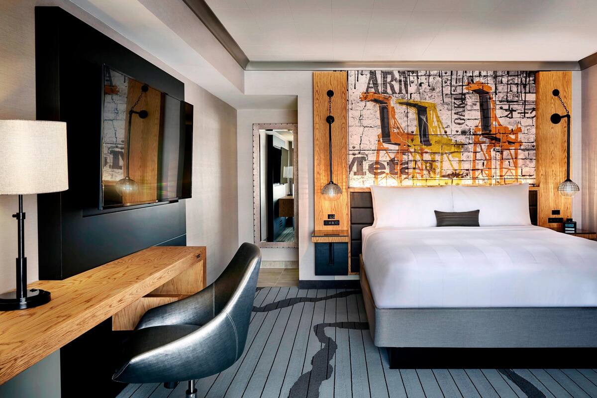 A modern hotel room with a large bed, a desk, a chair, and a unique wall art featuring construction vehicles.