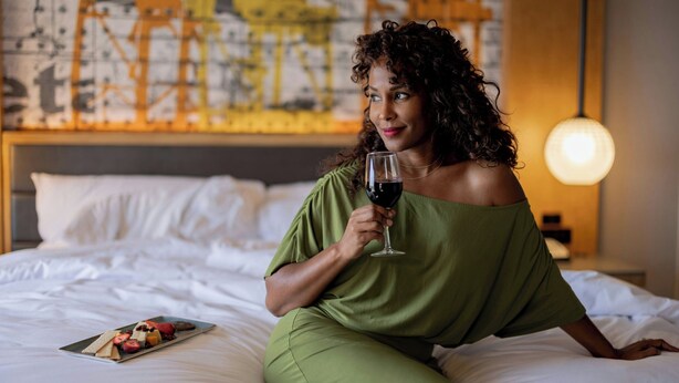 A person sitting on a bed with a plate of fruit and holding a glass of wine. - Oakland Marriott City Center (Oakland)