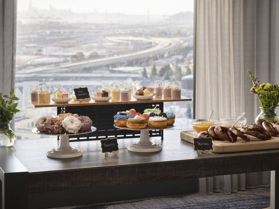 A buffet table with various desserts and snacks, a view of a cityscape through a window, and a vase with flowers on the left side.