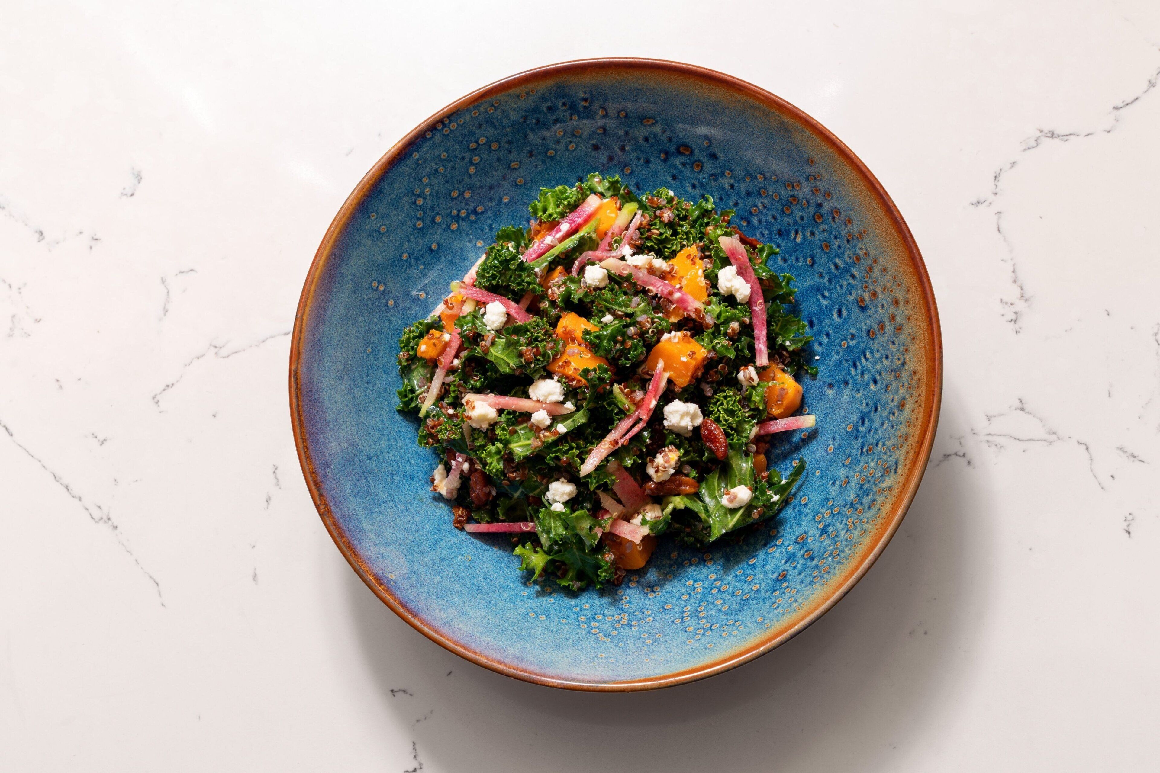 A blue ceramic bowl containing a salad with various vegetables and cheese on a white marble surface.