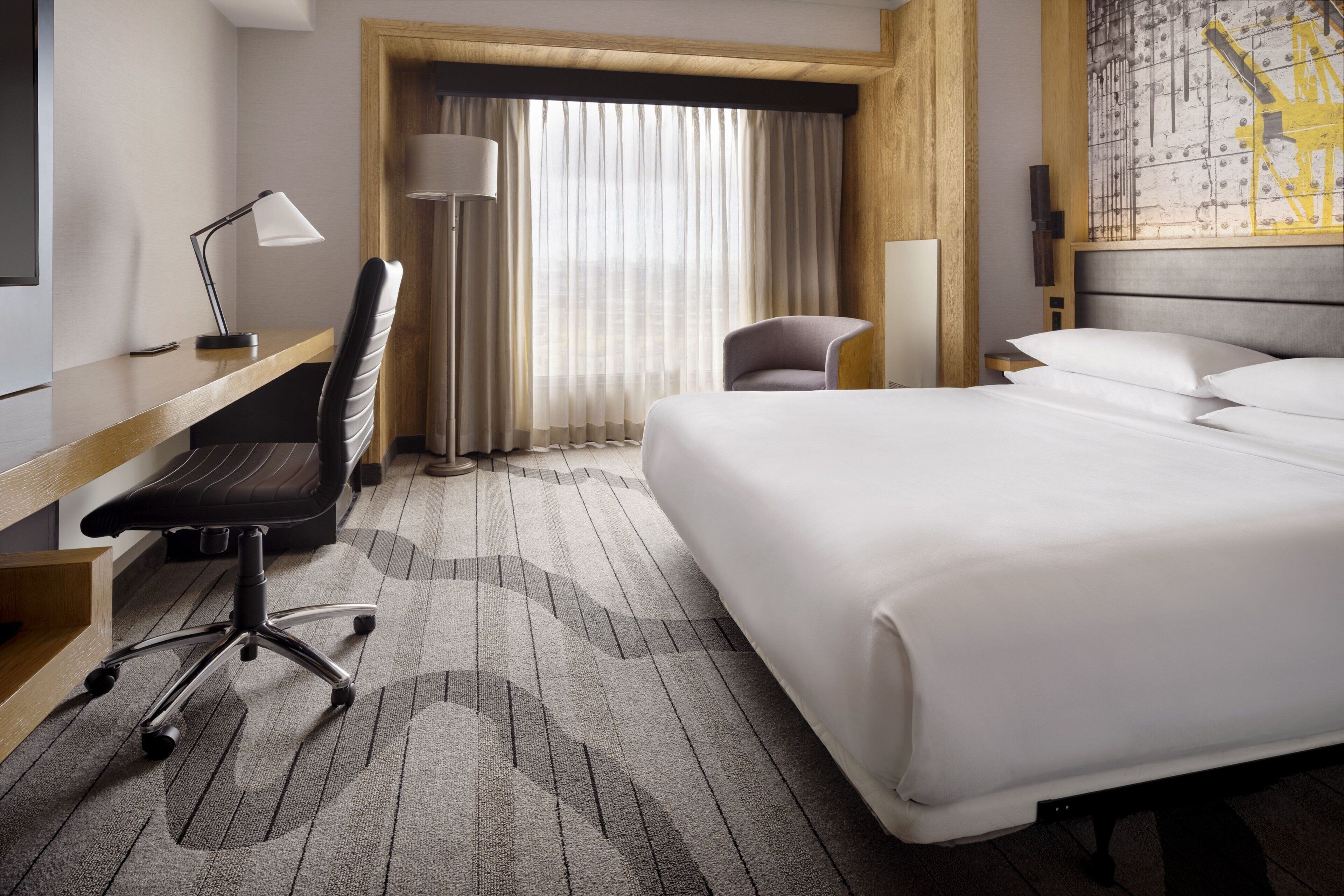 Habitación de hotel con cama, escritorio, silla y un ventanal con cortinas.