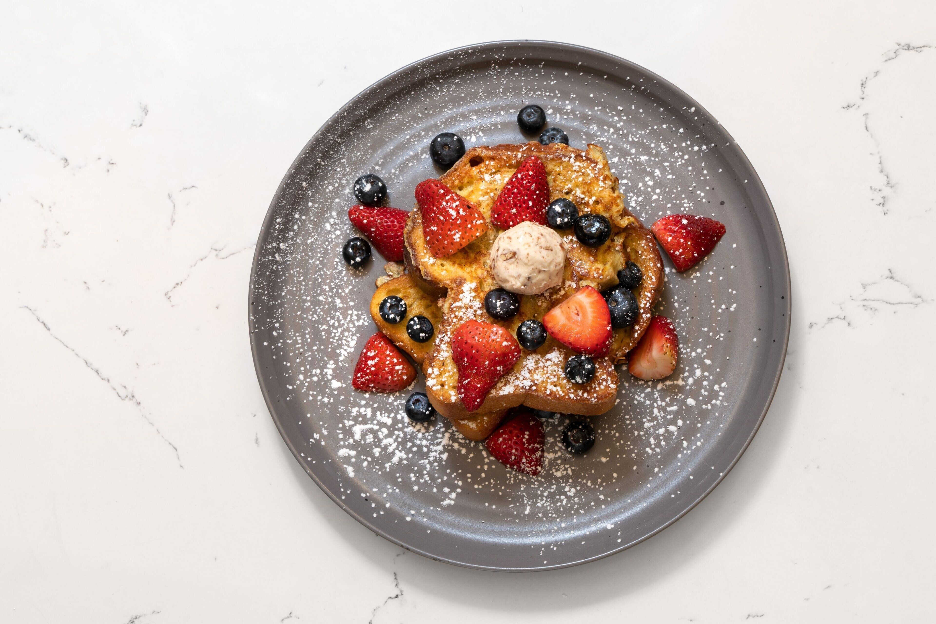 A plate of French toast with strawberries, blueberries, and a scoop of ice cream, dusted with powdered sugar.