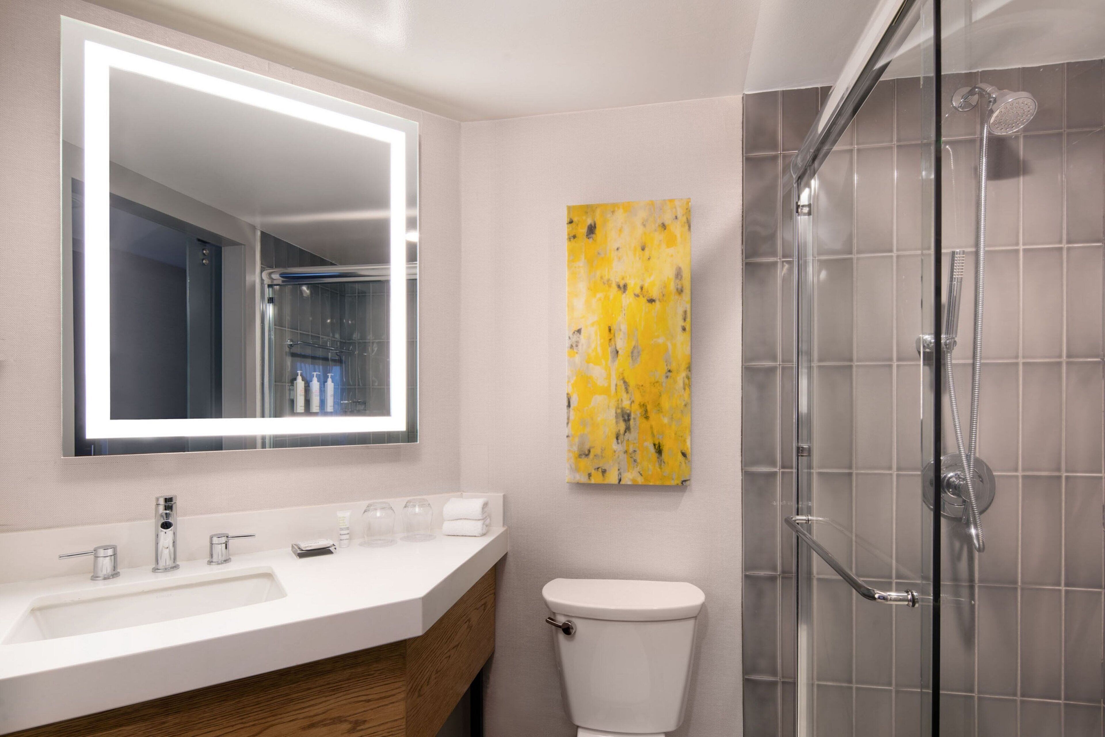 A modern bathroom with a white sink, a large mirror, a glass shower enclosure, and a yellow abstract painting on the wall.
