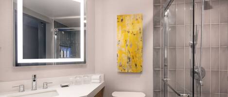 A modern bathroom with a white sink, a large mirror, a glass shower enclosure, and a yellow abstract painting on the wall.