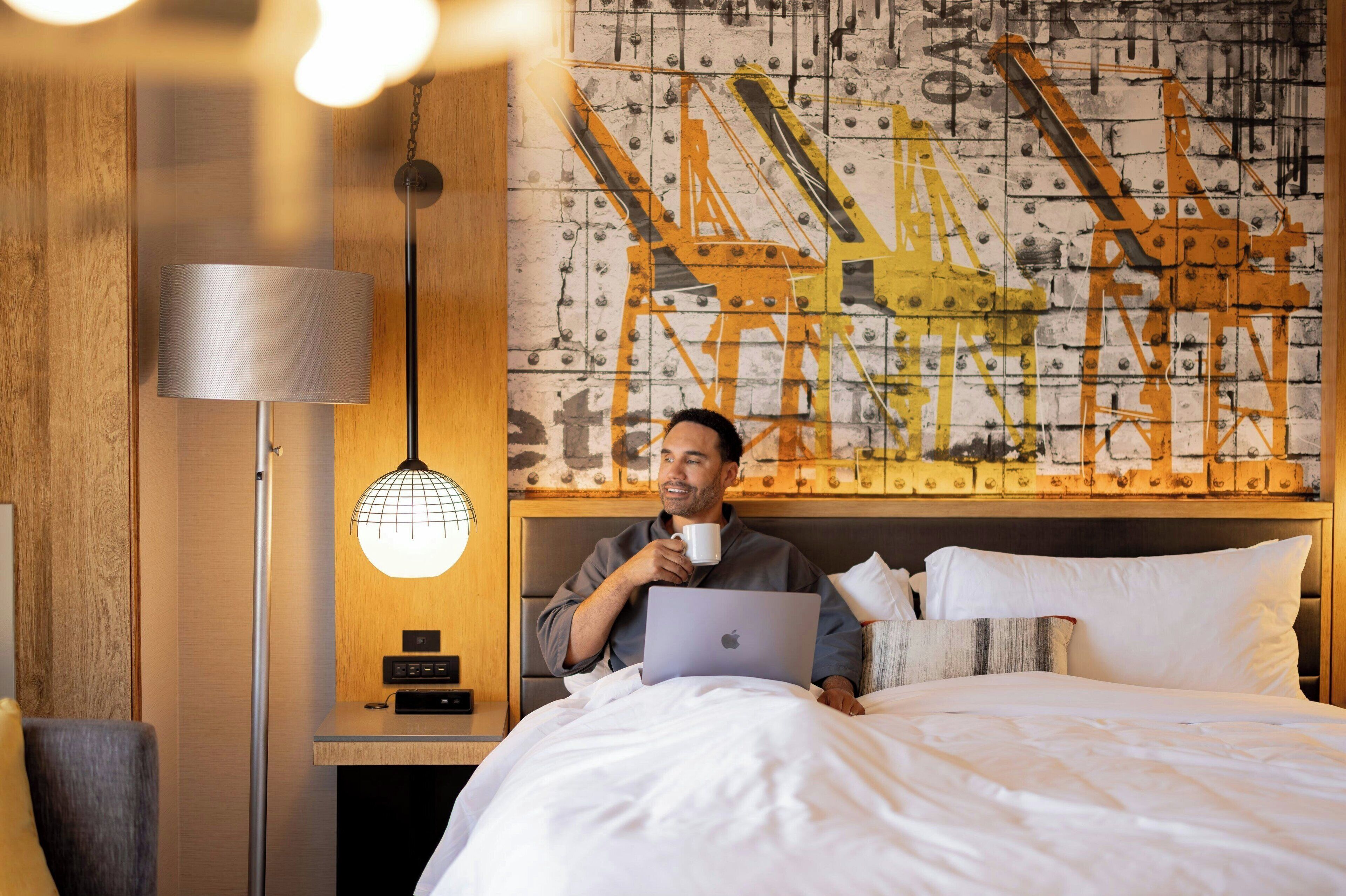 A hotel room with a bed, a lamp, a laptop, and a wall with a construction-themed mural.