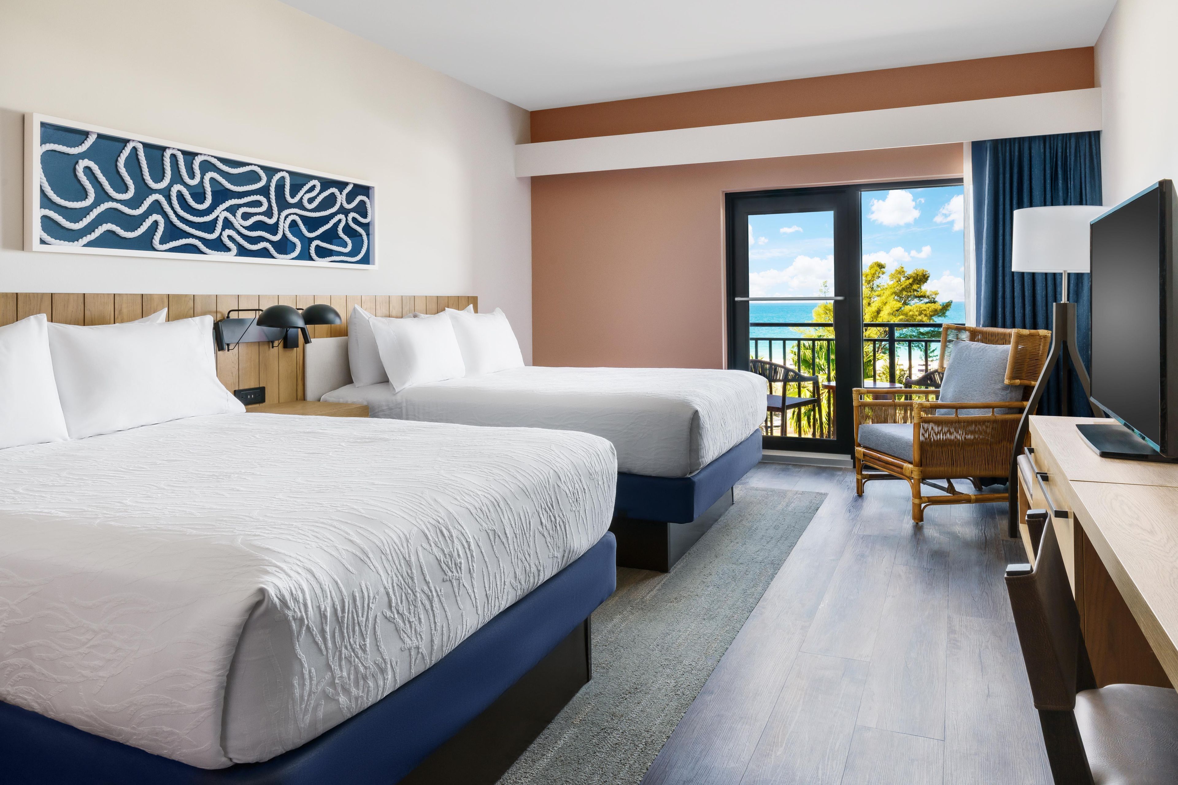 Habitación de hotel con dos camas, un escritorio, una silla, un televisor y un balcón con vista al mar.