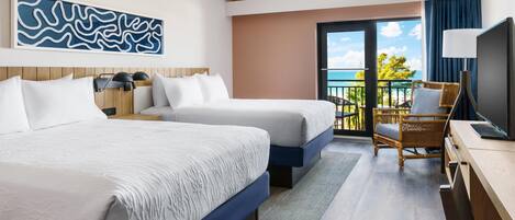 Habitación de hotel con dos camas, un escritorio, una silla, un televisor y un balcón con vista al mar.