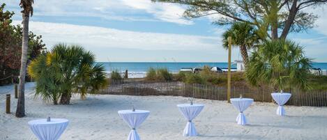 A beachside setting with four white tables set up on the sand, palm trees, and a fence in the background.