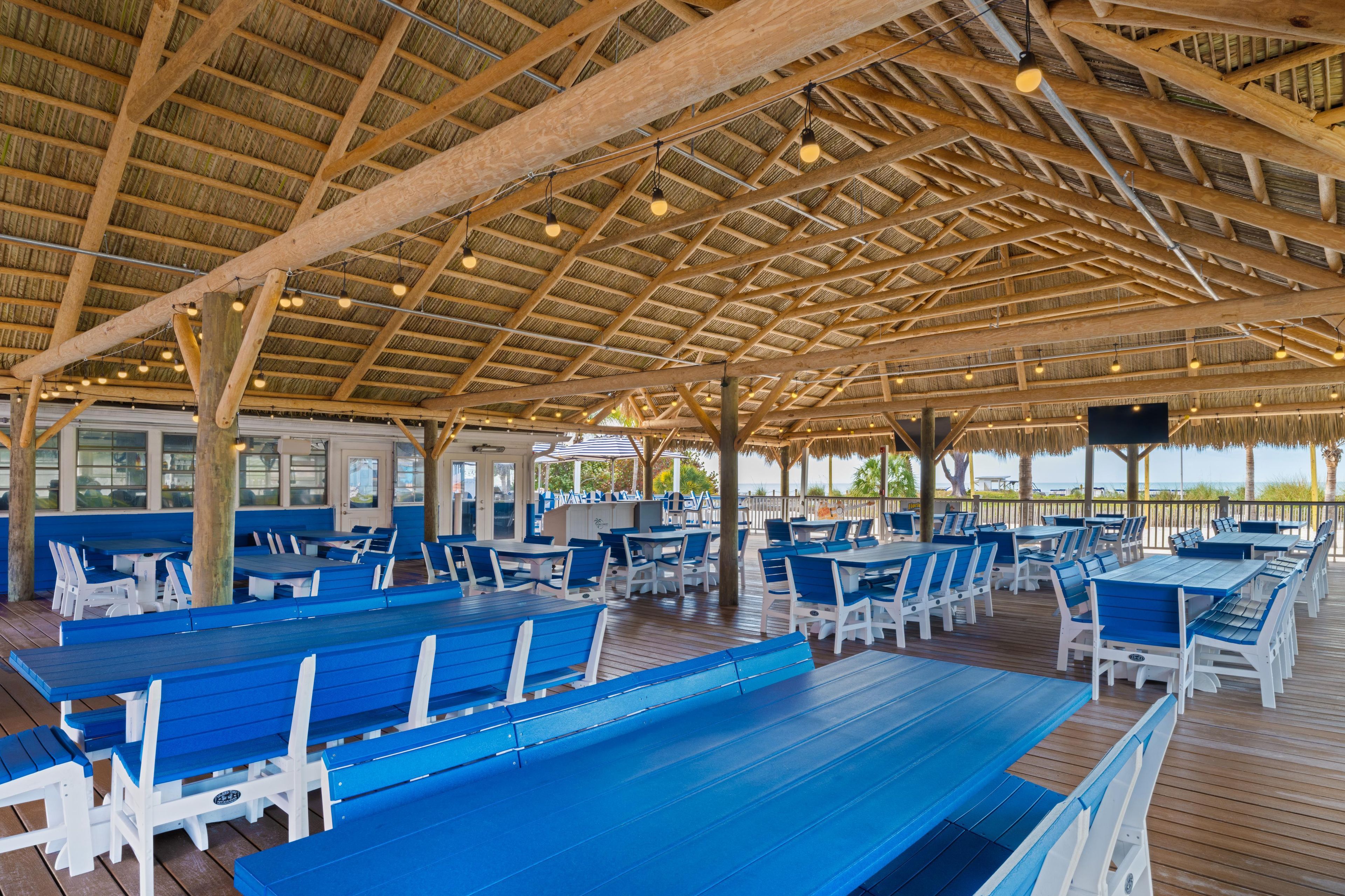 An outdoor dining area with wooden tables and blue chairs, under a thatched roof with exposed beams.
