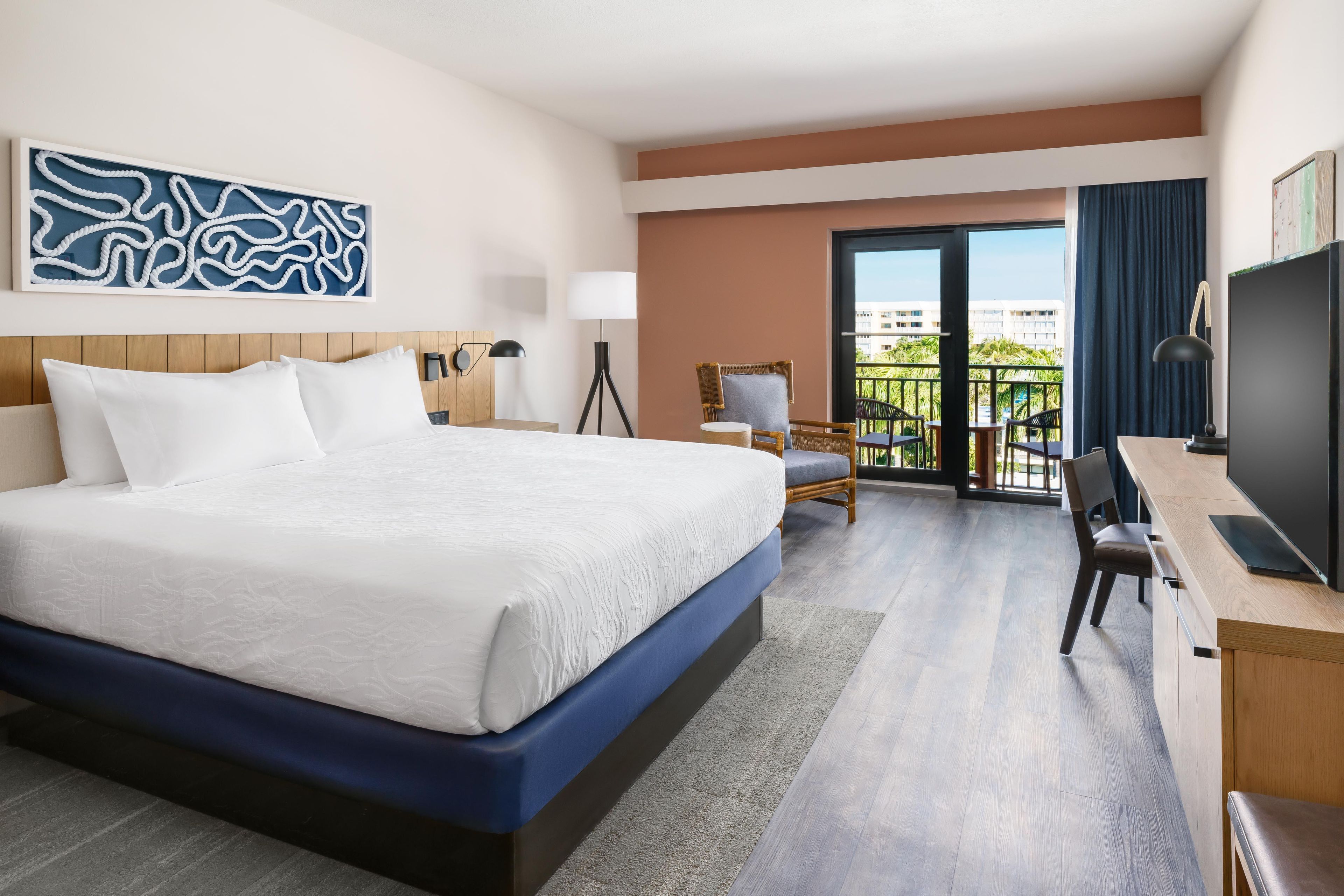 A hotel room with a large bed, a desk with a TV, a chair, a lamp, and a view of the city through the balcony.