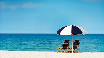 Two beach chairs under a striped umbrella on a sandy beach with a clear blue sea in the background.