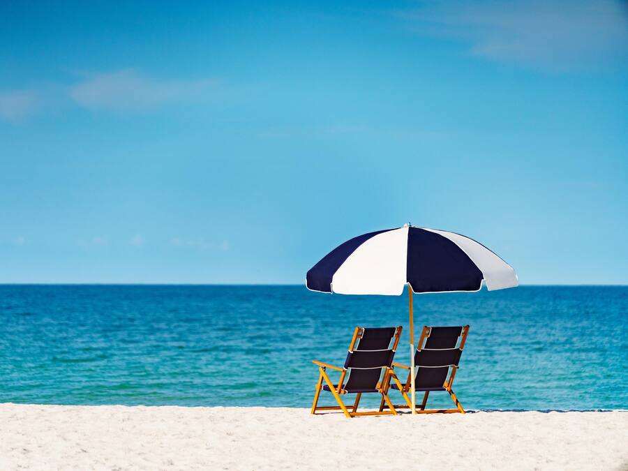 Two beach chairs under a striped umbrella on a sandy beach with a clear blue sea in the background.