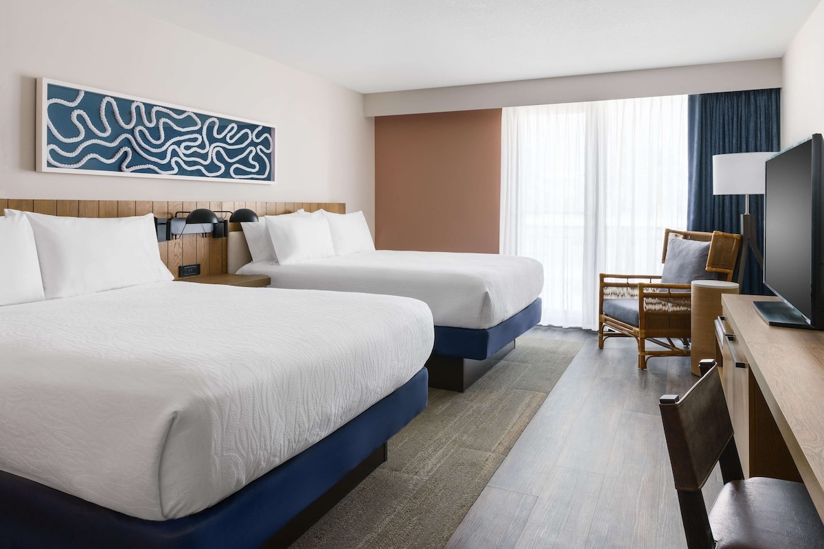 Habitación de hotel con dos camas, un escritorio, una silla, un televisor y un amplio ventanal con cortinas.