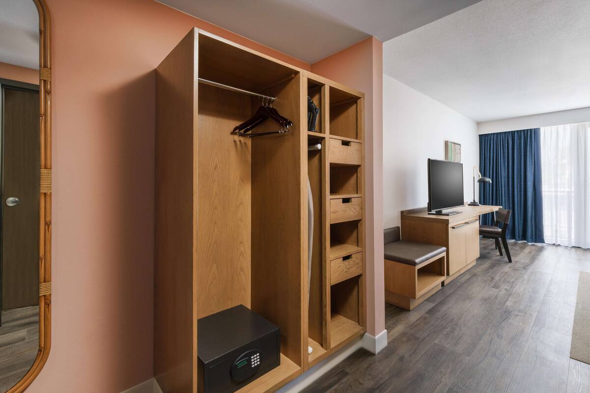 Une chambre d’hôtel avec une armoire en bois, un bureau avec un ordinateur, une télévision et une grande fenêtre avec des rideaux.