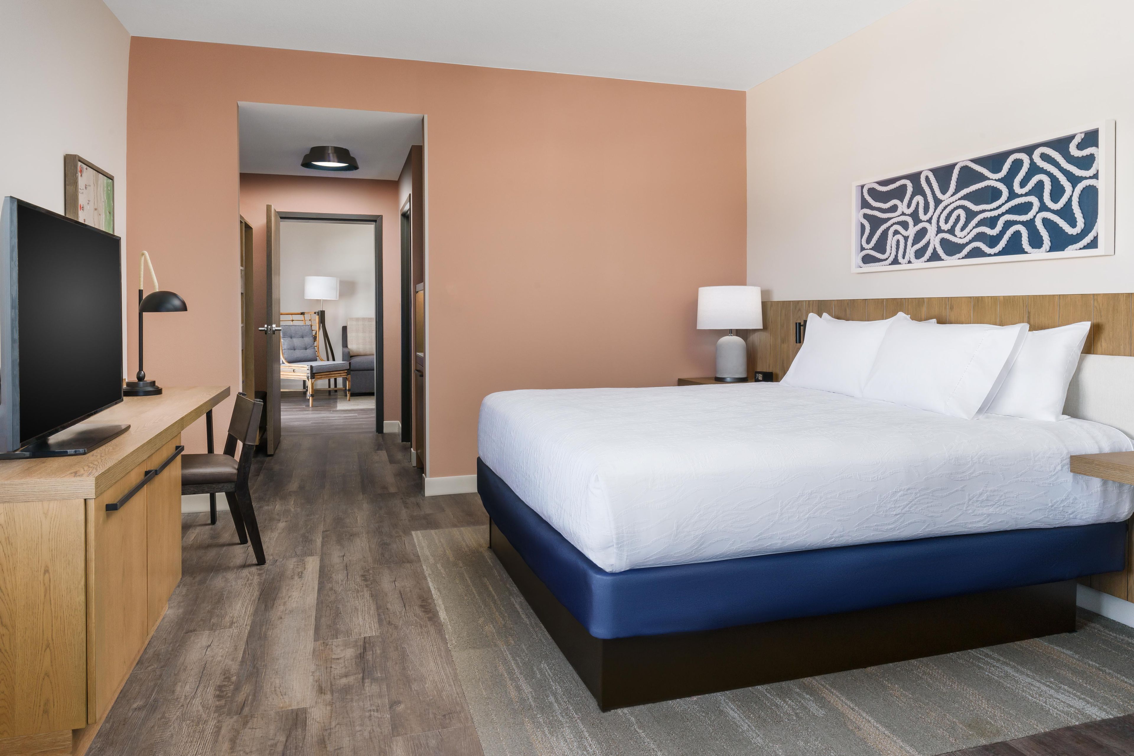 A hotel room with a bed, a desk, a television, and a hallway leading to another room.