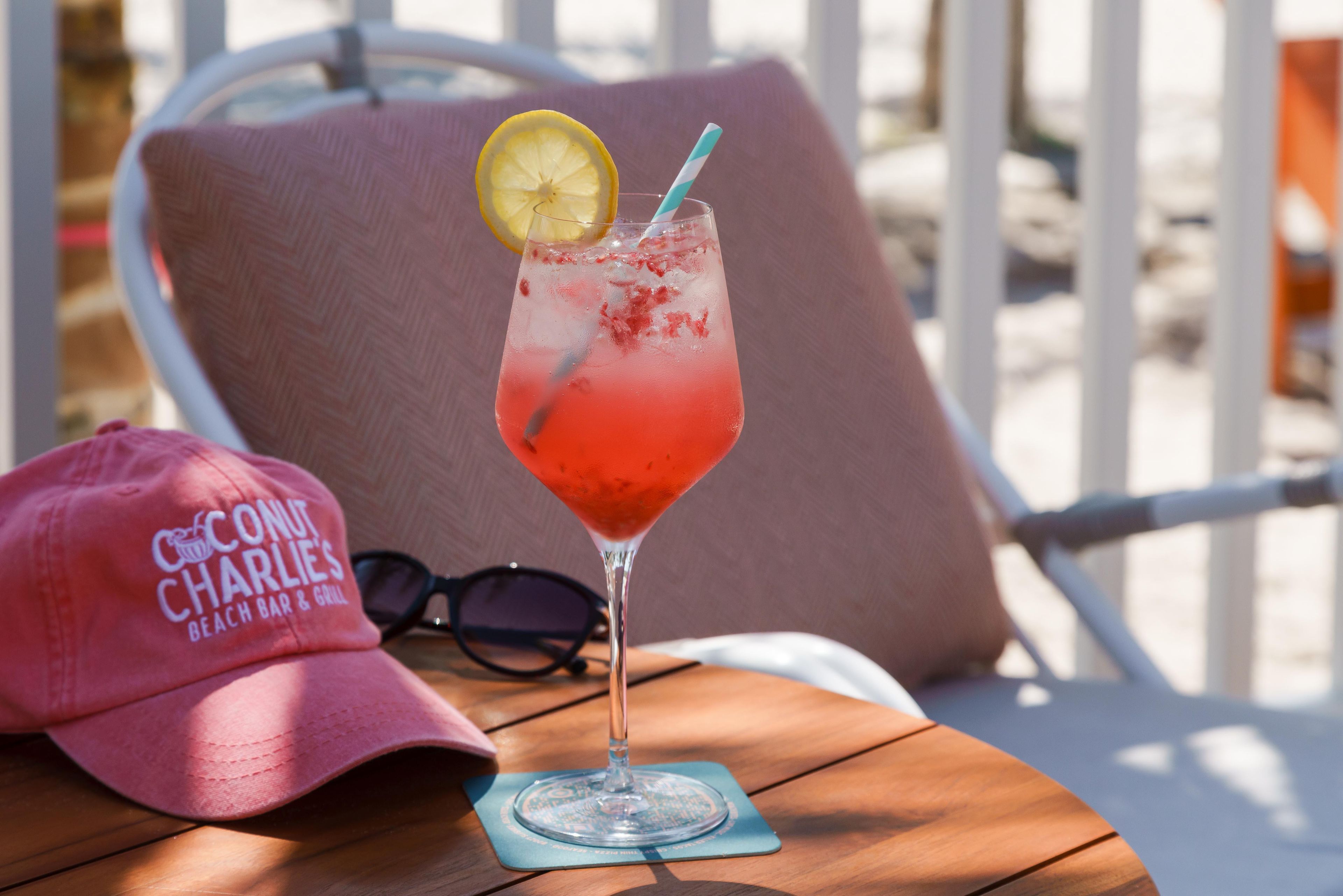 A cocktail glass with a lemon slice and a straw on a coaster, a pink cap with text, and a pair of sunglasses on a wooden table.
