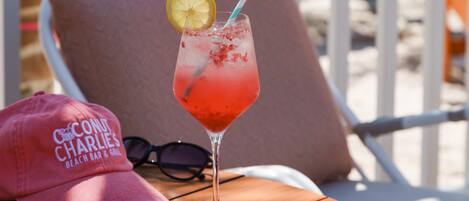 A cocktail glass with a lemon slice and a straw on a coaster, a pink cap with text, and a pair of sunglasses on a wooden table.
