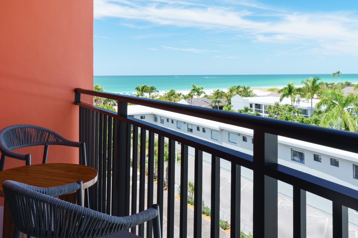 A balcony with a view of a beach, a table, and two chairs.