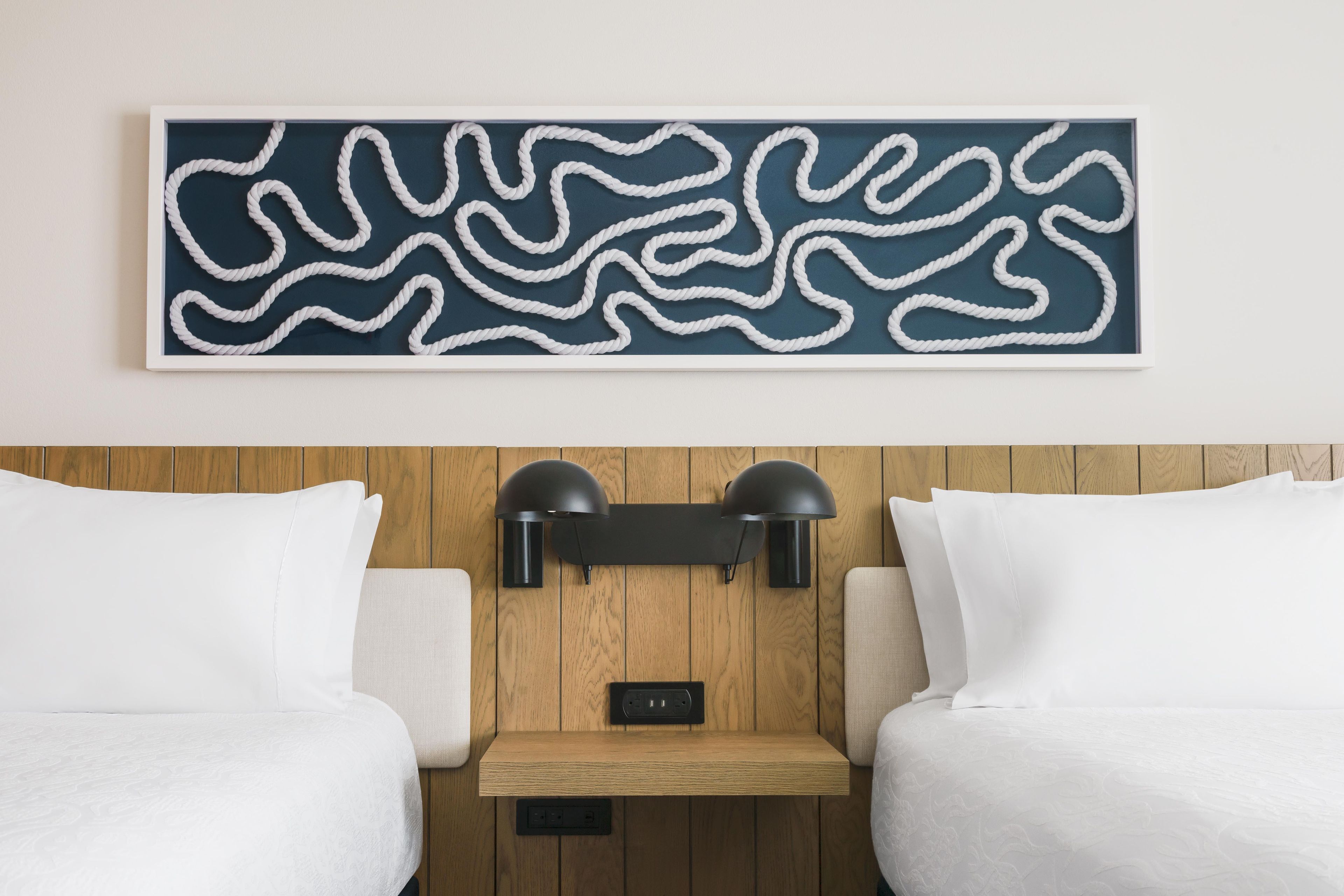 Habitación de hotel con un cuadro abstracto de gran tamaño, dos almohadas blancas, un cabecero de madera y una lámpara negra fijada a la pared.