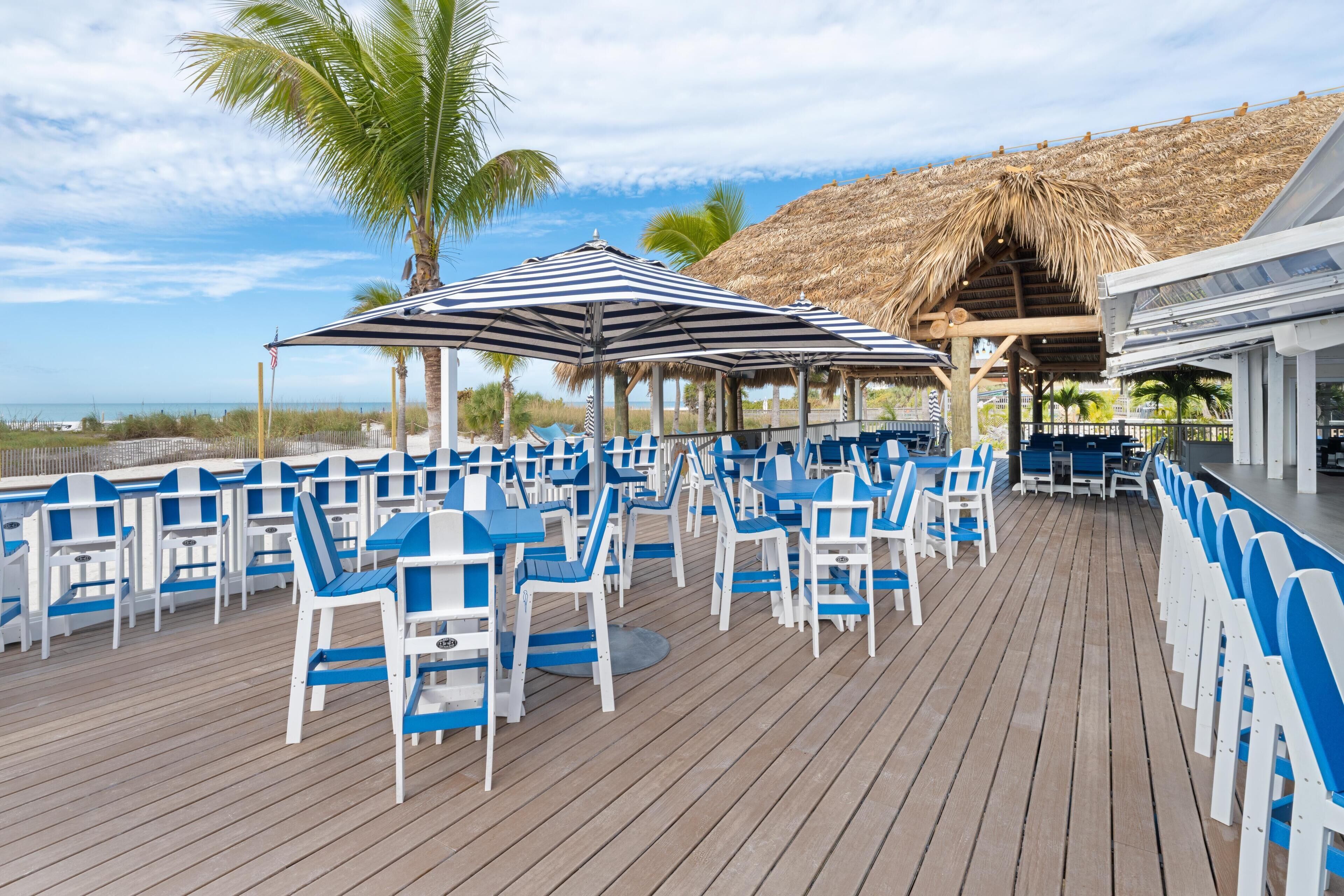 A deck with blue and white chairs and tables, a thatched roof structure, and a view of the ocean.