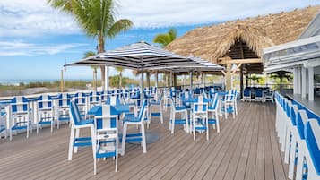 A deck with blue and white chairs and tables, a thatched roof structure, and a view of the ocean.