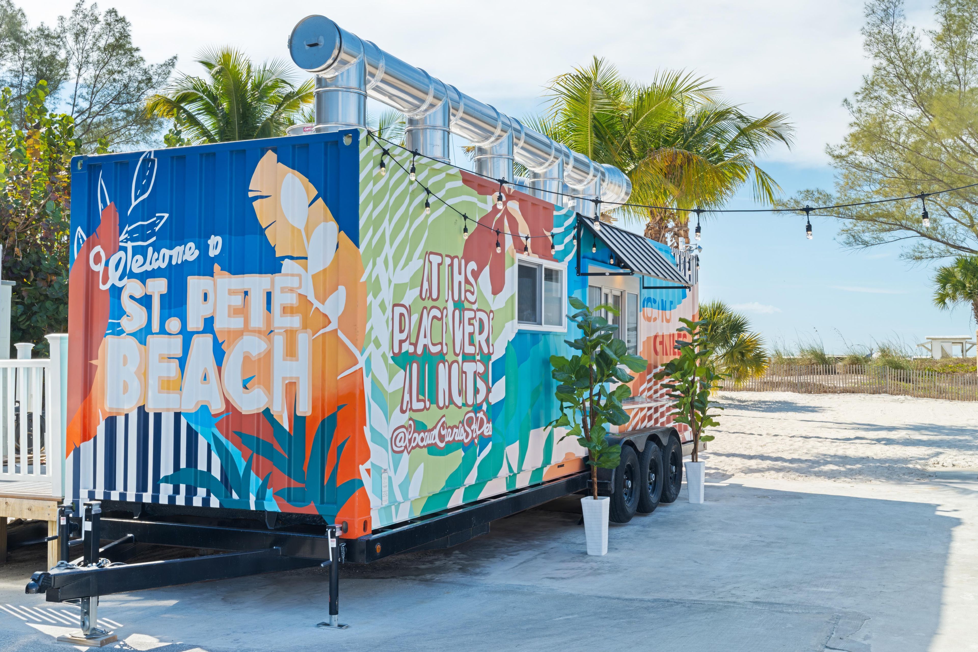 Um food truck colorido com temática tropical, estacionado numa área arenosa com palmeiras ao fundo.