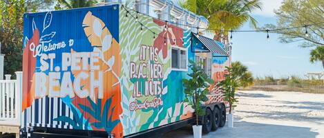 A colorful food truck with a tropical theme, parked on a sandy area with palm trees in the background.