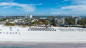 Vue aérienne d’un complexe hôtelier situé en bord de mer, avec plusieurs bâtiments, une plage de sable et un ciel bleu dégagé.