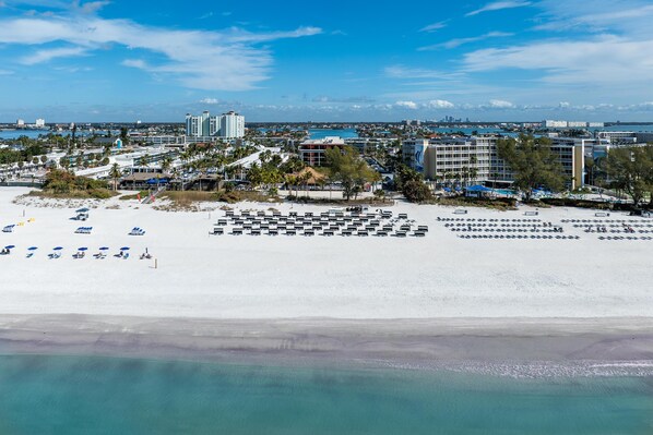 Ett flygbildsfoto av ett semesterområde vid stranden med flera hotellbyggnader, en sandstrand och en klarblå himmel.