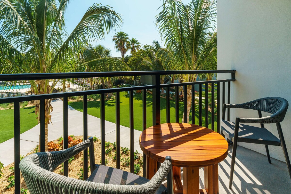 Un balcón con una mesa de madera y dos sillas de mimbre, con vista a una piscina y palmeras.
