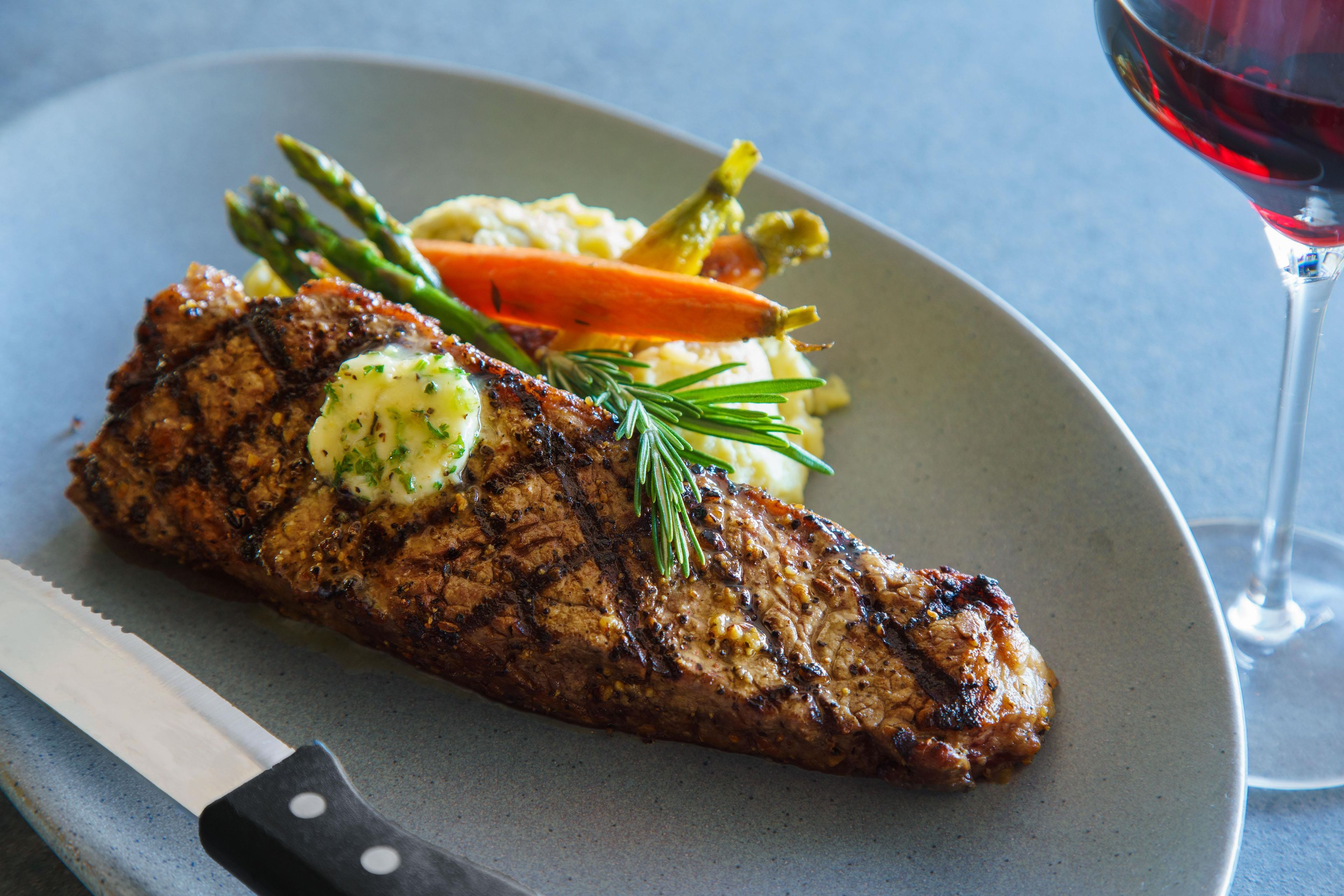A steak with a pat of butter and herbs, accompanied by vegetables and a glass of red wine on a plate.
