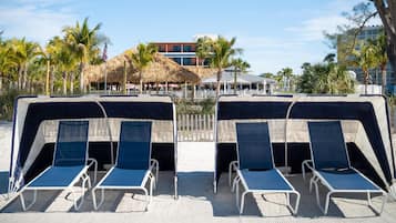 Two beach cabanas with blue cushions and white metal frames, a thatched-roof structure, and a building with balconies in the background.