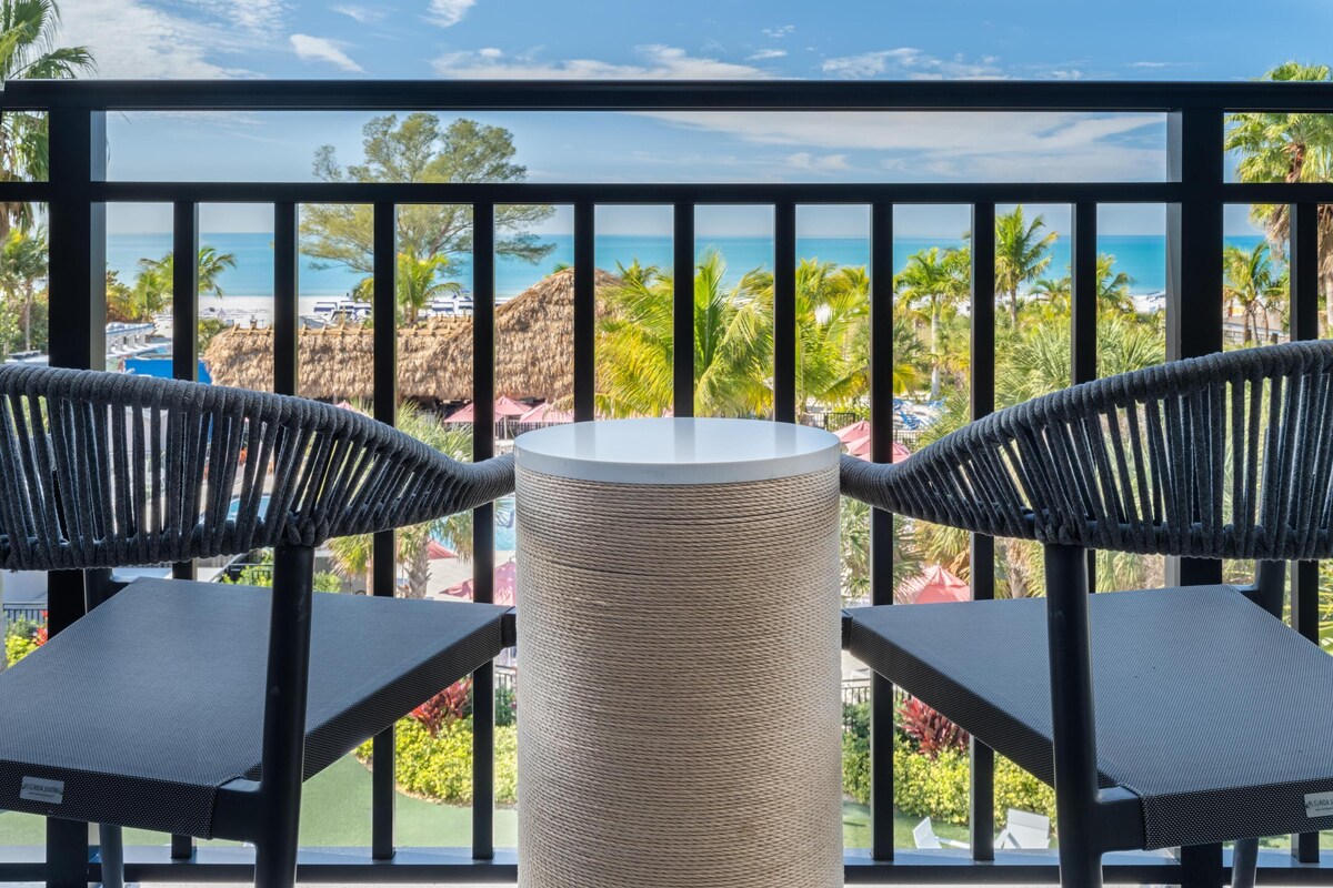 Un balcón con dos sillas de mimbre y una mesa redonda, con vistas a una playa con palmeras y un cielo azul despejado.