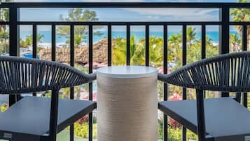 A balcony with two wicker chairs and a round table, overlooking a beach with palm trees and a clear blue sky.