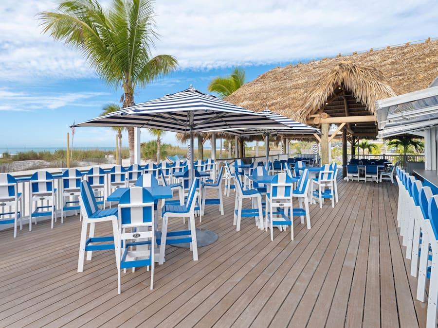 A deck with blue and white chairs and tables, a striped umbrella, and a thatched roof structure.