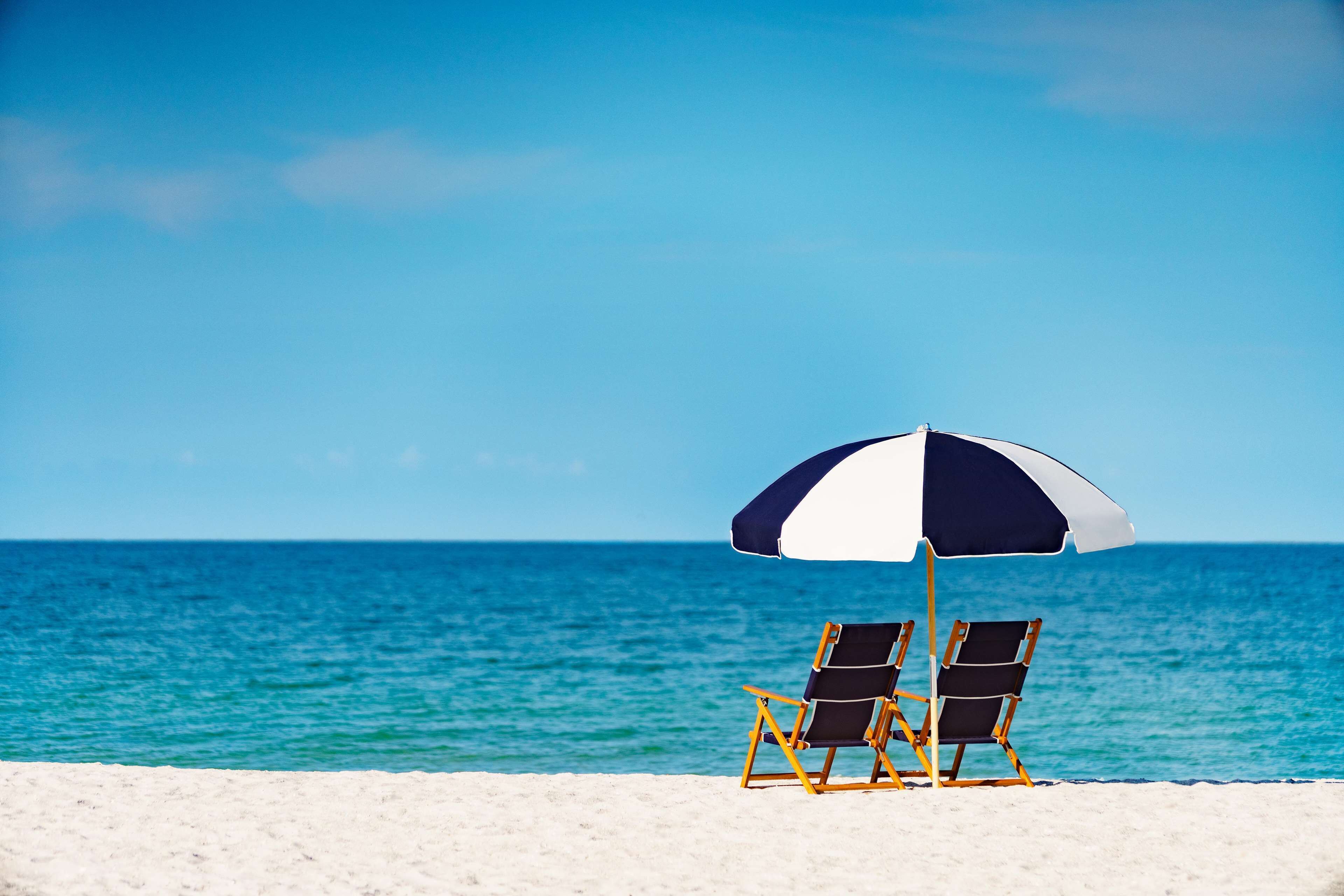 Zwei Liegestühle unter einem großen Sonnenschirm an einem sandigen Strand mit Blick auf das Meer.