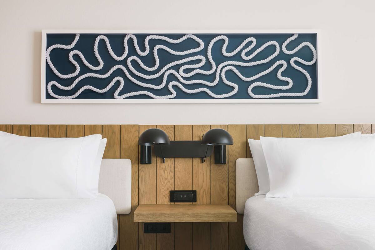 Une chambre d’hôtel avec une tête de lit en bois, deux oreillers blancs, une œuvre d’art abstraite encadrée et une étagère dotée d’une prise de courant.