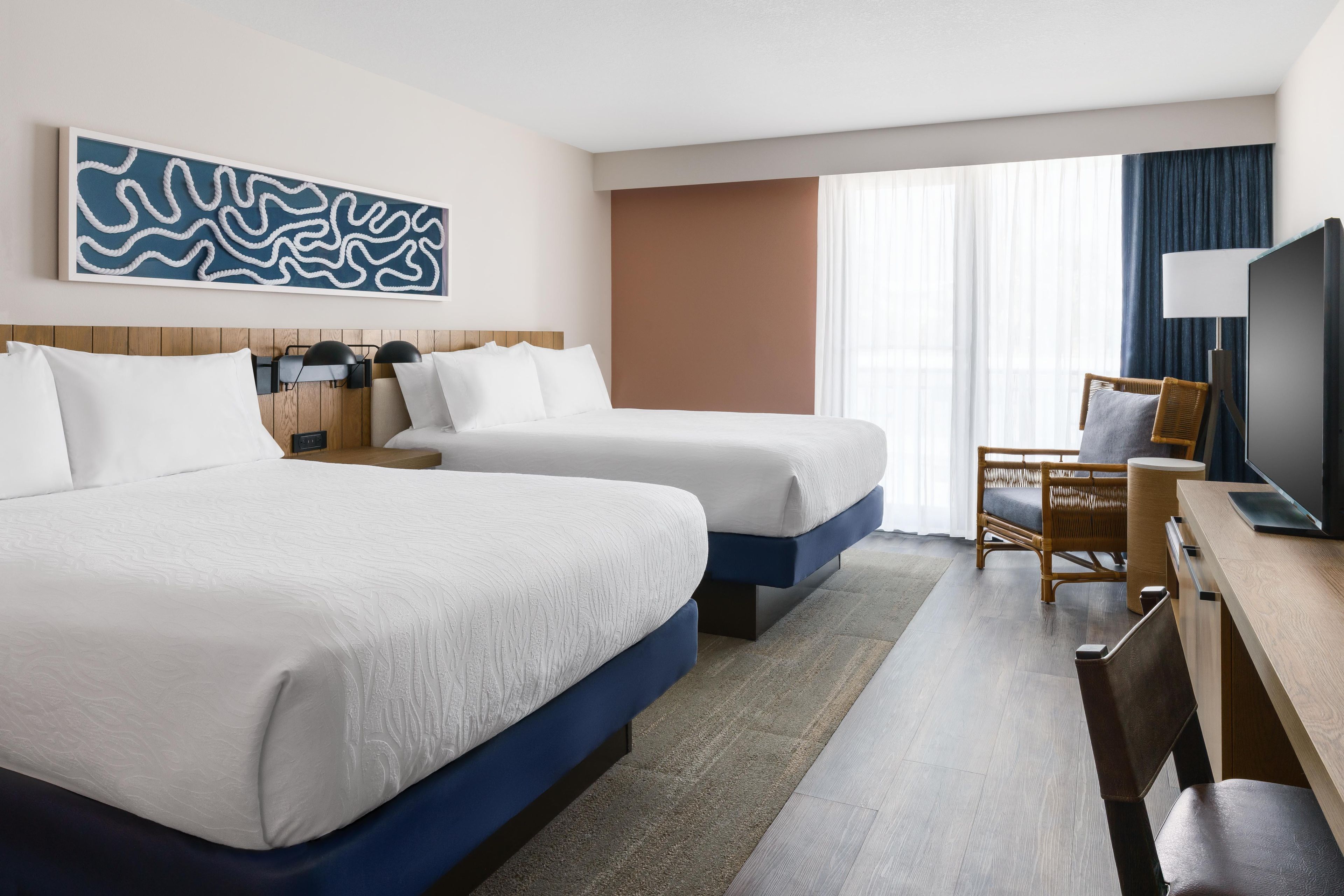 Ein Hotelzimmer mit zwei Betten, einem Schreibtisch, einem Stuhl, einem Fernseher und einem großen abstrakten Gemälde an der Wand.