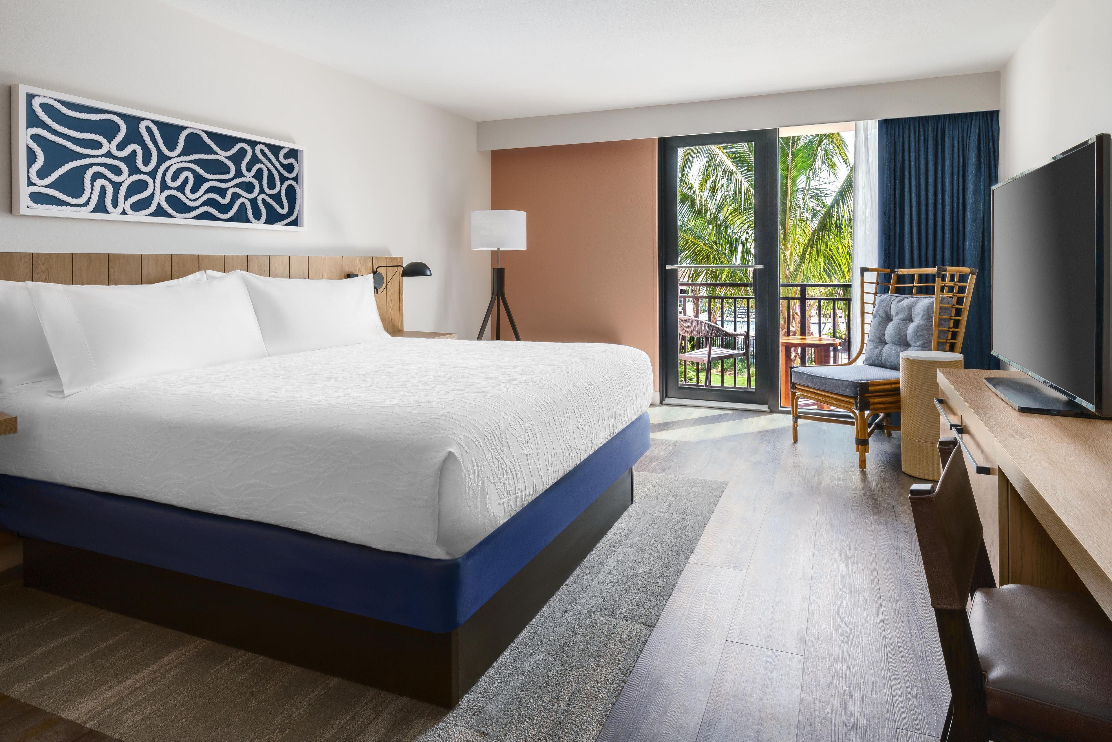 Une chambre d’hôtel avec un grand lit, un bureau avec une chaise, une télévision et un balcon offrant une vue sur les palmiers.