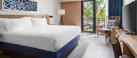 Ein Hotelzimmer mit einem großen Bett, einem Schreibtisch mit Stuhl, einem Fernseher und einem Balkon mit Blick auf Palmen.