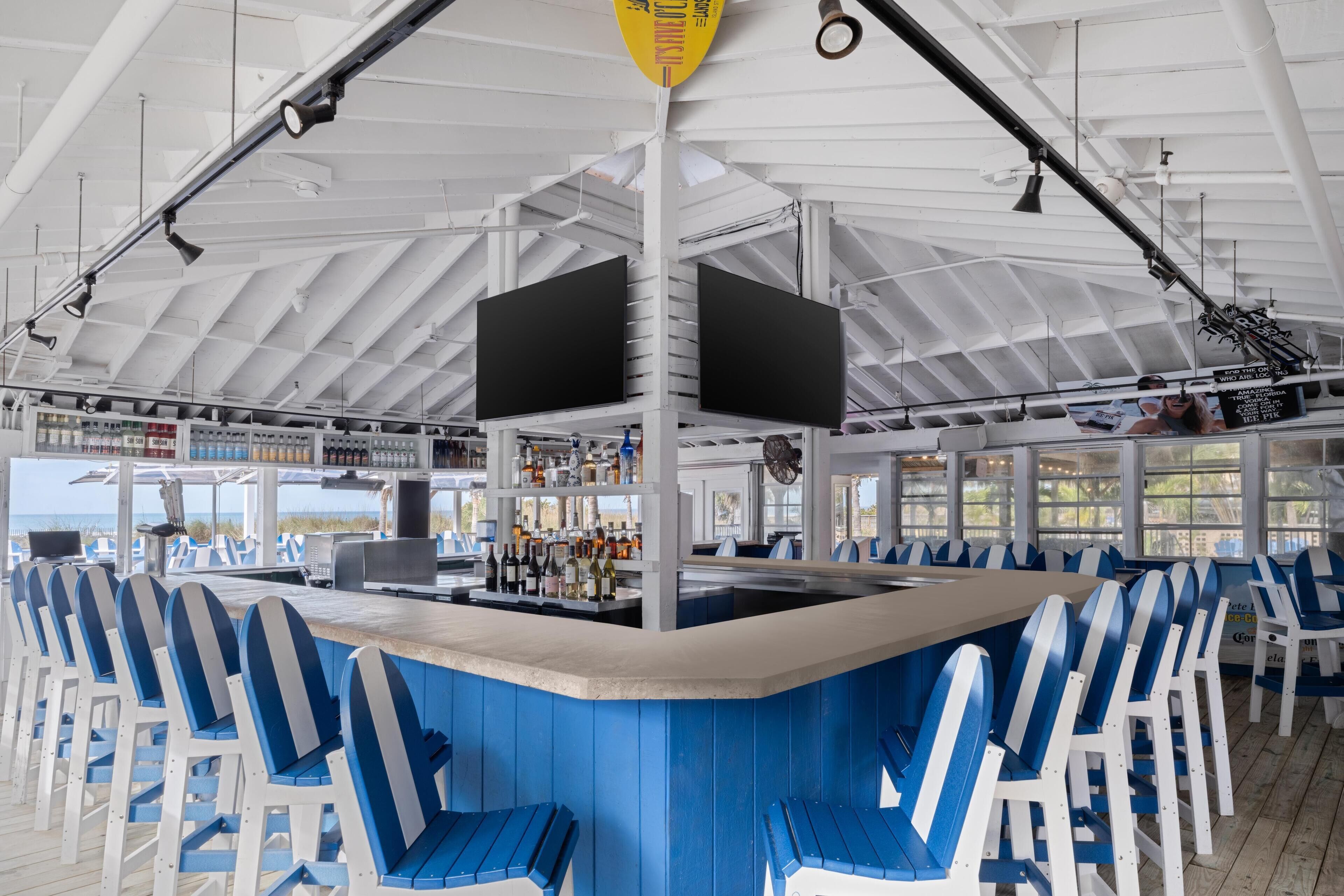 A bar area with blue and white chairs, a counter with a bar, and two large screens mounted on the ceiling.
