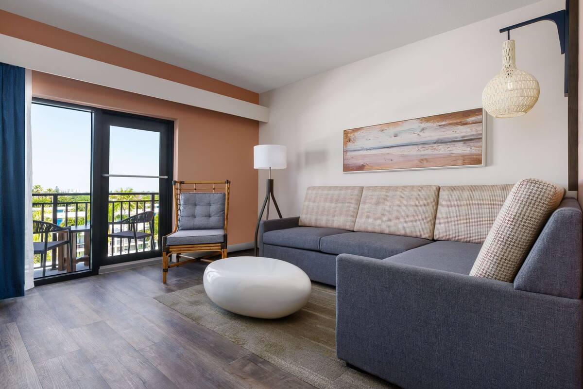Un salon moderne avec un canapé gris, un pouf blanc et un fauteuil. Une grande fenêtre offre une vue sur la verdure et donne accès à un balcon.