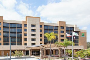A multi-story hotel with a modern architectural design, featuring a flat roof, large windows, and a landscaped entrance area with palm trees.