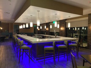 A modern bar area with a central island, bar stools, pendant lighting, and a view through large windows. - Holiday Inn Diamond Bar - Pomona by IHG (Diamond Bar)