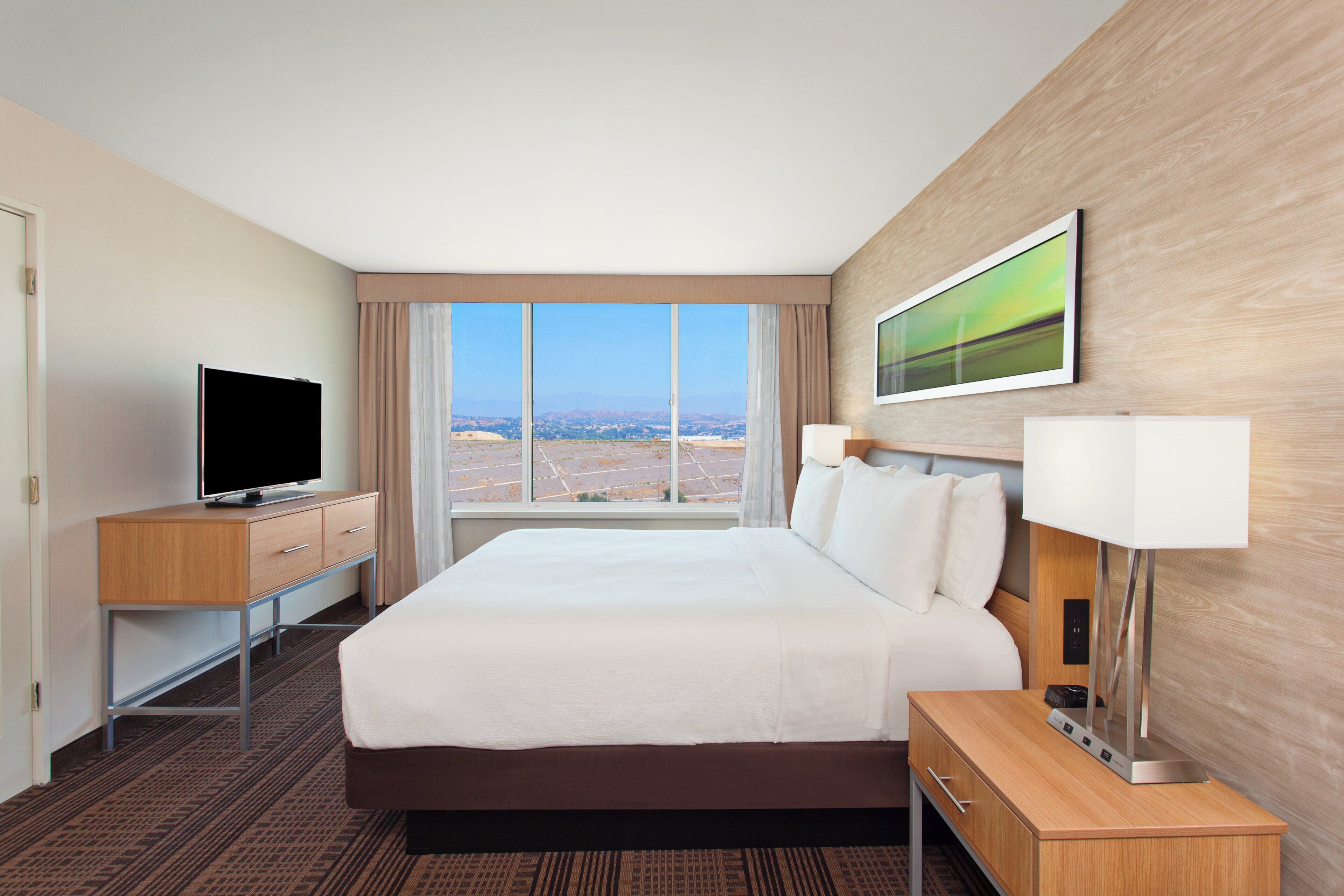 A hotel room with a large bed, a television on a wooden desk, a window with a view of a landscape, and a painting on the wall.
