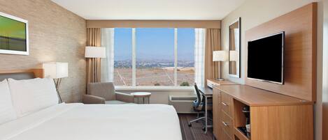 A hotel room with a large bed, a desk with a flat-screen TV, a chair, a small table, and a window with a view of the city.