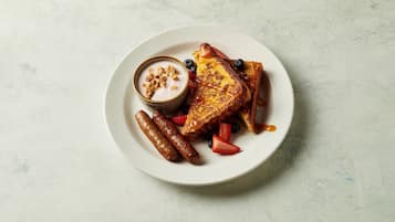 Un plato con tostadas francesas, salchichas, fresas, arándanos y un pequeño recipiente de yogur con granola.