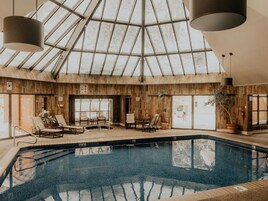 Una piscina interior con techo de vidrio, paredes de madera y sillas de descanso.
