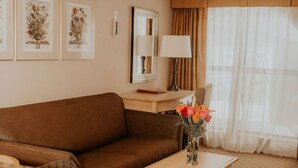 Habitación de hotel con un sofá marrón, un sillón estampado, una mesita de madera con un jarrón de flores, un escritorio con una lámpara e impresiones botánicas enmarcadas en la pared.