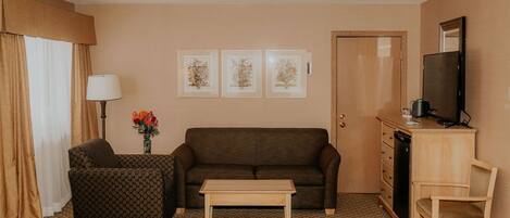 A hotel room with a brown sofa, a patterned armchair, a wooden coffee table, a TV on a wooden stand, and a door with a mirror above it.